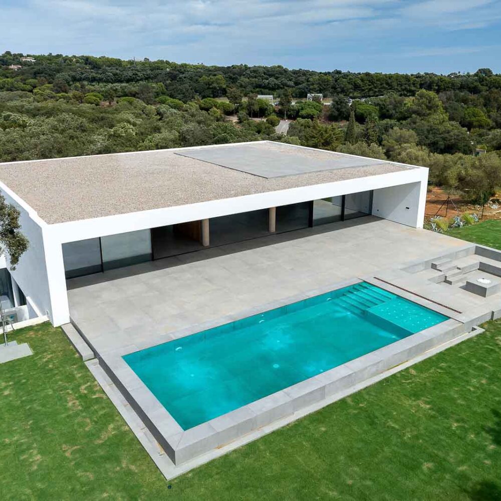 Aerial view of modern white Villa 93, with a pool, patio, integrated outdoor lounge, and green lawn among trees.