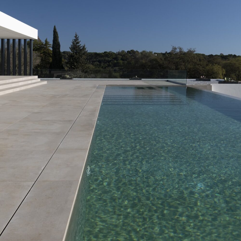 Modern villa with a large patio, steps, an infinity pool, and a backdrop of lush green trees under a blue sky.