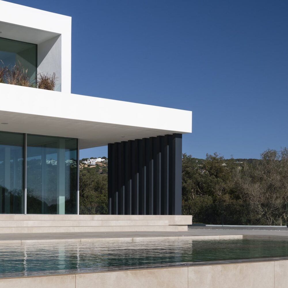 Modern luxury villa in Sotogrande with large glass windows, a dark slatted wall, an infinity pool, and green hills.