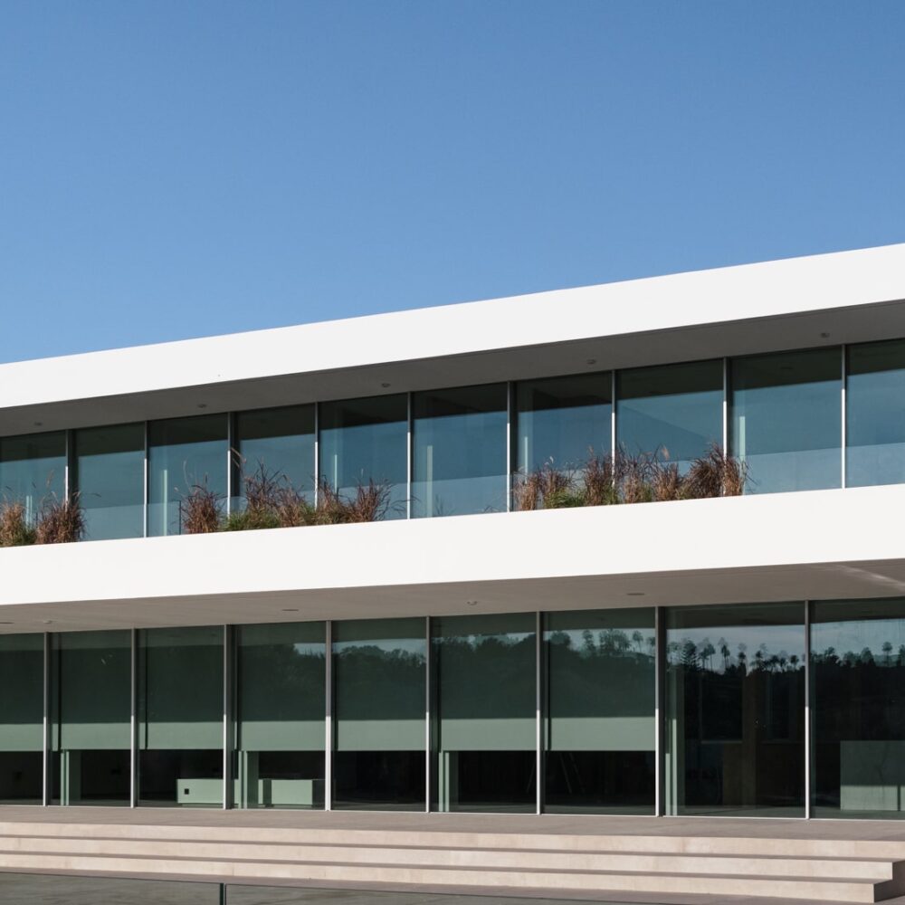 Modern two-story luxury villa with expansive glass windows and a balcony adorned with plants under a clear blue sky.