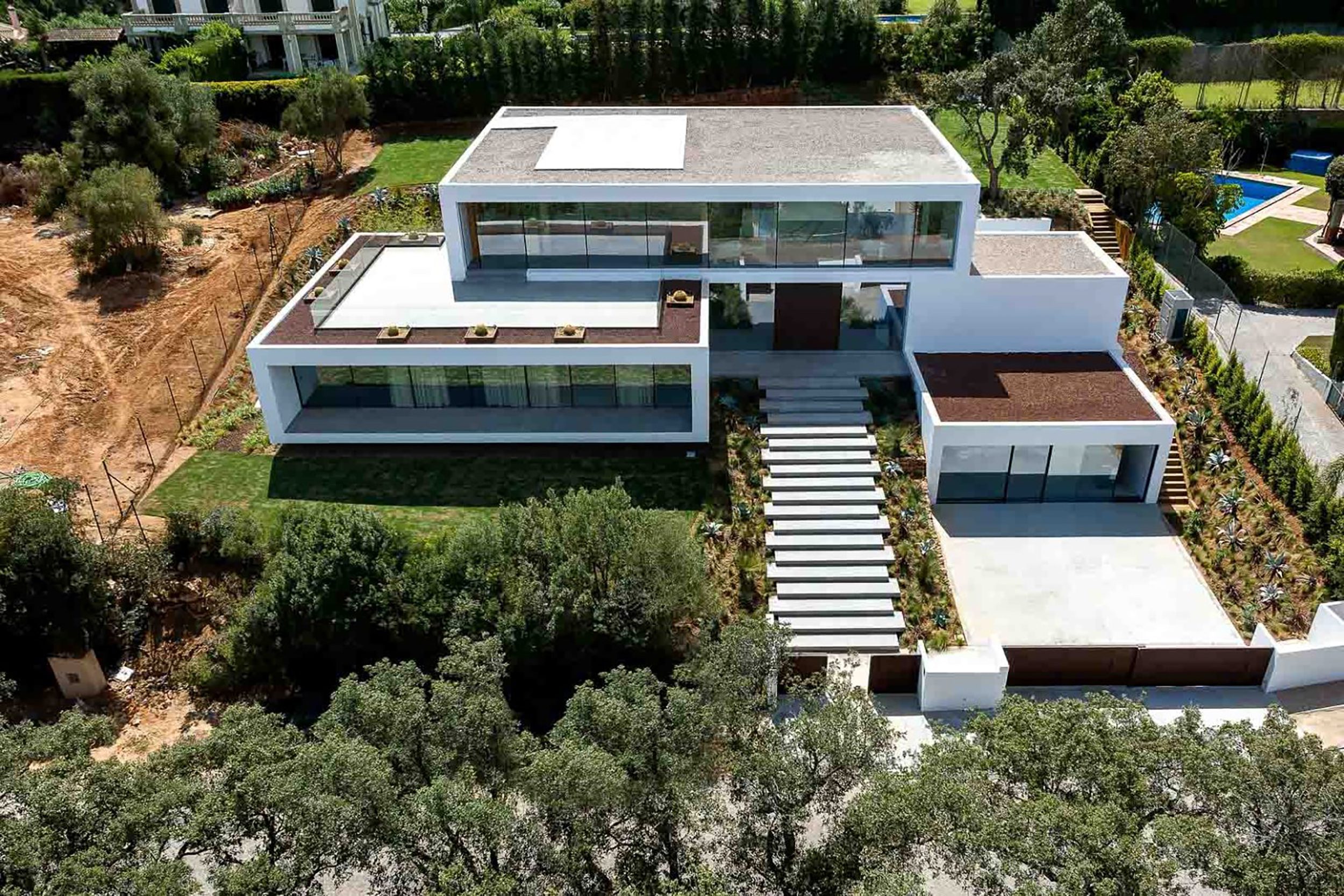 Vista aérea de la moderna Villa 93 con fachada blanca, grandes ventanales y escaleras en un entorno verde de Sotogrande.