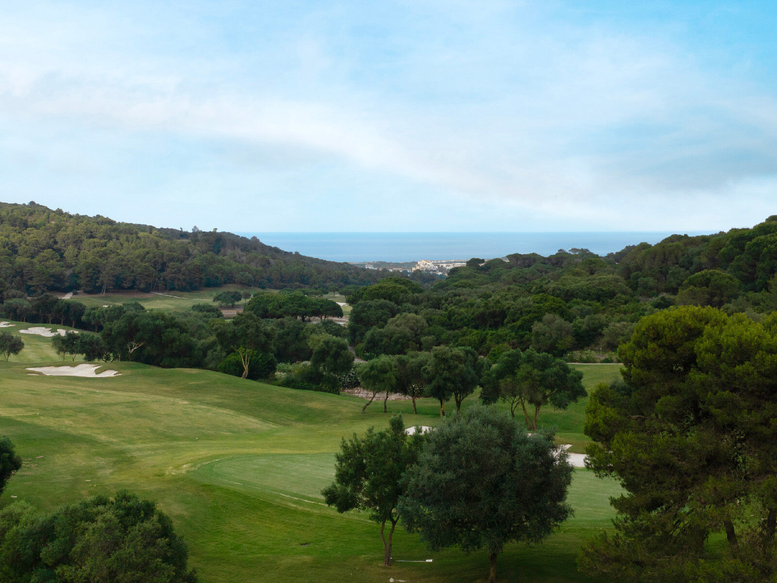 Verdant Sotogrande golf course with trees and bunkers, overlooking the sea and a distant coastal town.