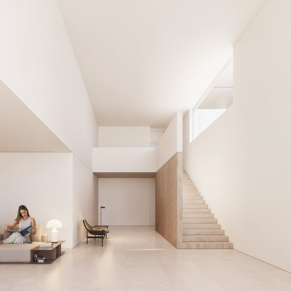 Woman reads in a bright, minimalist living room of a luxury villa with a large staircase.